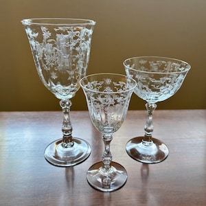 May include: Three clear glass goblets with etched floral designs. The goblets vary in size, with a tall wine glass, a smaller stemmed glass, and a coupe glass. The glasses are displayed on a wooden surface.