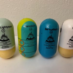May include: Four colorful, capsule-shaped Disney character toys. The toys are labeled "Buzz Lightyear", "Heimlich", "Sebastian", and "Lucky", with corresponding numbers. Each has a castle graphic and the text "Happiest Celebration on Earth".