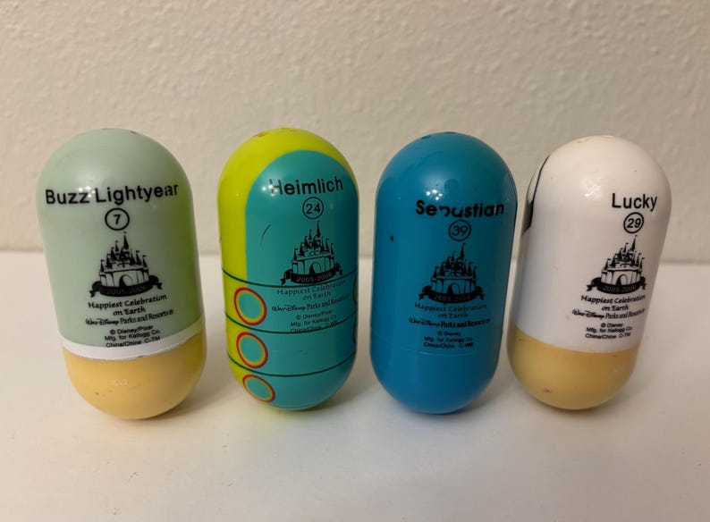 May include: Four colorful, capsule-shaped Disney character toys. The toys are labeled "Buzz Lightyear", "Heimlich", "Sebastian", and "Lucky", with corresponding numbers. Each has a castle graphic and the text "Happiest Celebration on Earth".