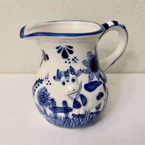 Könnte beinhalten: Weißer Keramikkrug mit blauen Blumen- und Kuhmotiven. Der Krug hat einen Ausguss, einen Griff mit blauen Streifen und ein detailliertes Relief einer Kuh auf der Vorderseite. Der Boden und der Rand sind ebenfalls blau eingefasst, was einen klassischen, dekorativen Look erzeugt.