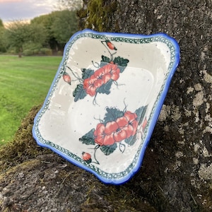 Puede incluir: Un plato cuadrado de cerámica con un diseño floral. El plato tiene un interior color crema con flores rojas y hojas verdes. El borde es azul con un borde festoneado. El plato está sobre un fondo natural.