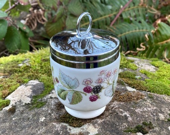 Könnte beinhalten: Ein weißer Eierbecher aus Keramik mit einem silbernen Deckel und einem Blumendesign mit Brombeeren und Blättern. Der Eierbecher hat einen kleinen Griff auf dem Deckel.