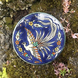 May include: A round, hand-painted ceramic plate with a vibrant bird design. The plate features a deep blue background with a white bird, orange, yellow, and green accents, and floral motifs. The plate has a turquoise rim.