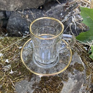 Può includere: Un set di tazza e piattino in vetro trasparente con finiture dorate. La tazza ha un manico e un design a coste. Il piattino è rotondo e ha anche un bordo dorato. Il set è appoggiato su una roccia muschiosa.