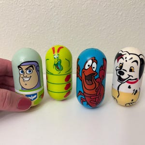 May include: Four colorful, egg-shaped toys featuring cartoon characters. One has Buzz Lightyear's face, another a green caterpillar-like creature, a red crab, and a Dalmatian dog. The toys are on a white surface.