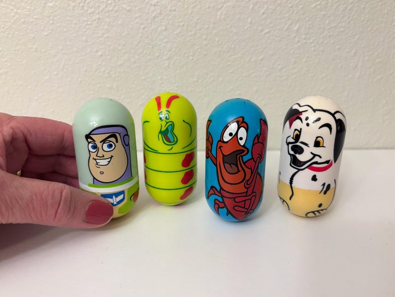May include: Four colorful, egg-shaped toys featuring cartoon characters. One has Buzz Lightyear's face, another a green caterpillar-like creature, a red crab, and a Dalmatian dog. The toys are on a white surface.