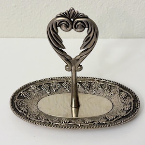May include: Silver-toned, two-tiered serving tray with an oval base and a decorative handle. The base features a grape and leaf design around the edge. The handle is heart-shaped.