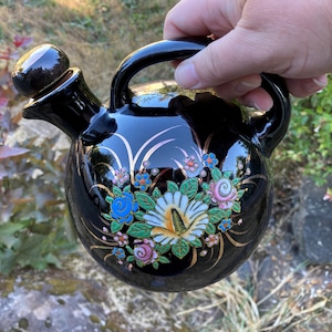 Pode incluir: Decantador de licor de cerâmica preta com um design floral dourado e uma rolha preta. O decantador tem um corpo redondo e uma alça.