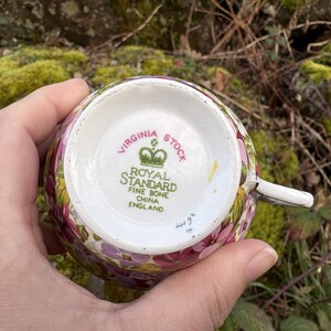 May include: A floral teacup with the text "VIRGINIA STOCK" and "ROYAL STANDARD FINE BONE CHINA ENGLAND" on the bottom. The cup has a white handle and is decorated with pink, purple, and yellow flowers.