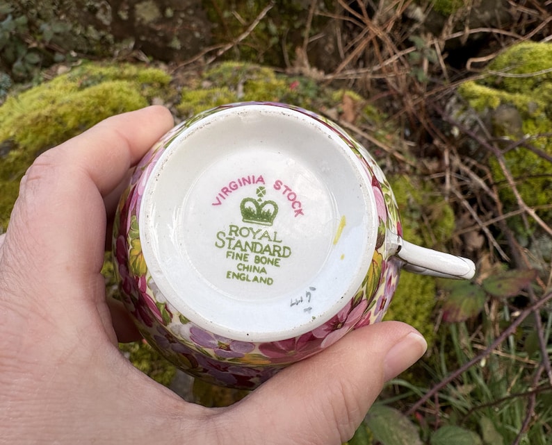 May include: A floral teacup with the text "VIRGINIA STOCK" and "ROYAL STANDARD FINE BONE CHINA ENGLAND" on the bottom. The cup has a white handle and is decorated with pink, purple, and yellow flowers.
