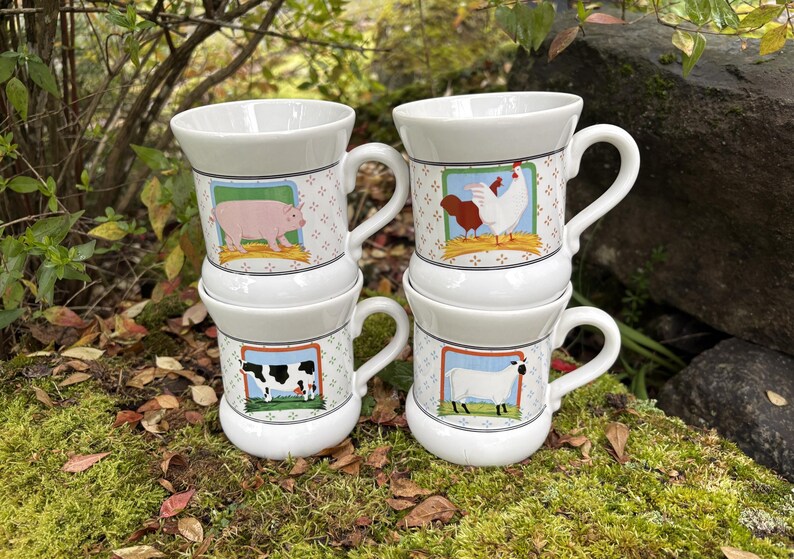 May include: Four white ceramic mugs stacked, each with a different farm animal illustration: a pig, a chicken, a cow, and a sheep. The mugs have a patterned border and a curved handle. The mugs are on a mossy surface.