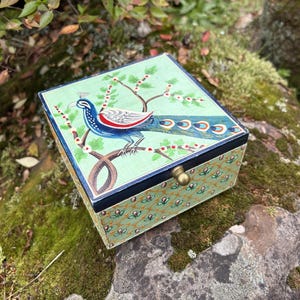 May include: A decorative box with a hinged lid, featuring a peacock design. The box has a light green background with a peacock illustration in blue, red, and white. The sides of the box are decorated with a floral pattern. A brass-coloured clasp secures the lid.