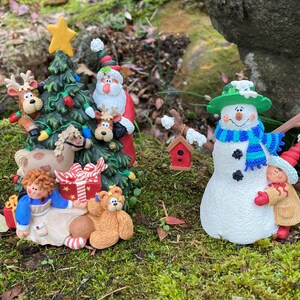 May include: A Christmas scene with a snowman, a Christmas tree, and various figurines. The snowman is wearing a green hat and a blue scarf. The Christmas tree is decorated with lights and ornaments. The figurines include a teddy bear, a reindeer, and a Santa Claus.