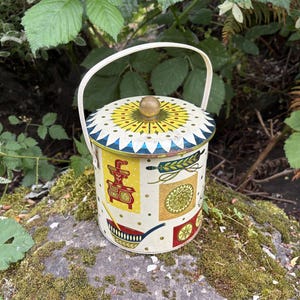 May include: A cylindrical metal tin with a cream-colored base, featuring a white handle and a lid topped with a gold-colored knob. The exterior is decorated with colorful illustrations of kitchen items, including a red stove and wheat stalks.