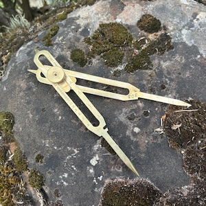May include: A brass drafting compass with two pointed legs and a central adjustment knob. The tool features engraved measurement markings along one arm and the number 305 stamped near the hinge. The metal surface has a metallic gold finish.