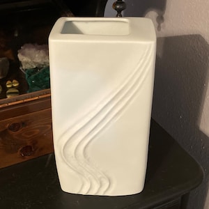 May include: White ceramic vase with a wavy, abstract design. The vase is tall and rectangular.