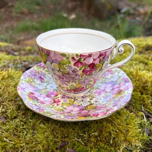 May include: A floral teacup and saucer set. The teacup and saucer are decorated with a pattern of pink, purple, and white flowers. The teacup has a gold rim and handle. The saucer has a scalloped edge.