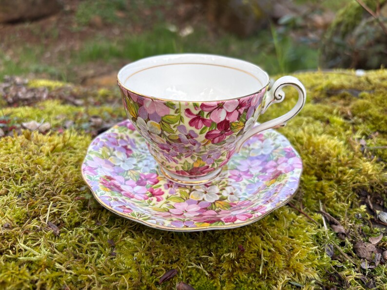 May include: A floral teacup and saucer set. The teacup and saucer are decorated with a pattern of pink, purple, and white flowers. The teacup has a gold rim and handle. The saucer has a scalloped edge.