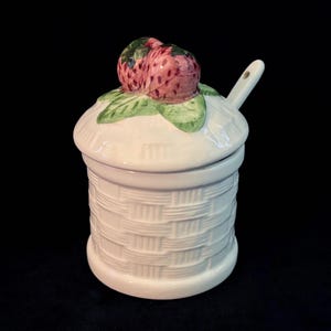 Könnte beinhalten: Ein weißes Keramikgefäß mit Deckel und Löffel. Das Gefäß hat ein Korbgeflecht-Design. Der Deckel ist mit zwei rosa Erdbeeren und grünen Blättern verziert. Der Löffel liegt auf dem Deckel. Der Hintergrund ist schwarz.
