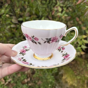May include: A pink teacup and saucer set with a floral rose pattern. The teacup has a gold rim and handle, and the saucer has a matching design. The set is held in front of a blurred green background.