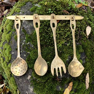 May include: Four antique brass kitchen utensils hanging on a brass bar. The set includes a slotted spoon, a solid spoon, a fork, and a ladle. The utensils are displayed against a moss-covered rock, with fallen leaves in the background.