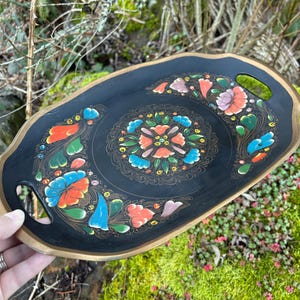 Könnte beinhalten: Schwarzes, ovales Dekotablett mit goldenem Rand und Griffen. Das Tablett hat ein Blumenmuster in Rot, Orange, Blau und Rosa. Das Tablett ist wahrscheinlich aus Holz oder einem ähnlichen Material.