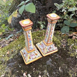 May include: Two square-based ceramic candle holders featuring a yellow background with hand-painted floral motifs, green vines, and small animal illustrations. The tapered columns have orange and brown trim at the top and bottom edges.