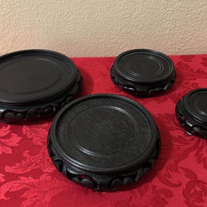 May include: A set of four circular black wooden display stands of varying sizes, each featuring a flat top surface and a carved, decorative base with scalloped edges. The stands are arranged in a group, showcasing their different diameters.