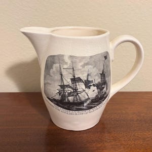 May include: Cream ceramic jug with a handle and spout. The jug has a black and white illustration of sailing ships and text. The jug is on a wooden surface.