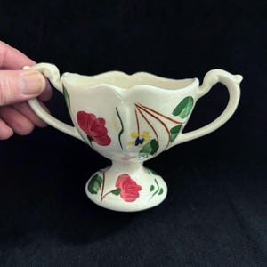 May include: White ceramic creamer or gravy boat with two handles. The boat has a scalloped edge and is decorated with hand-painted flowers in shades of red, green, yellow, and brown.