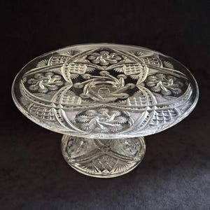 Puede incluir: Un soporte para pasteles de vidrio tallado transparente con una parte superior ovalada y una base de pedestal. El vidrio presenta intrincados patrones florales y geométricos. Diseñado para exhibir pasteles y otros postres.