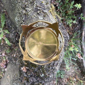 Puede incluir: Un portavelas de metal dorado con base circular y dise&ntilde;o calado. El portavelas tiene un patr&oacute;n de estrella en el borde superior. El portavelas est&aacute; sobre un tronco de &aacute;rbol.