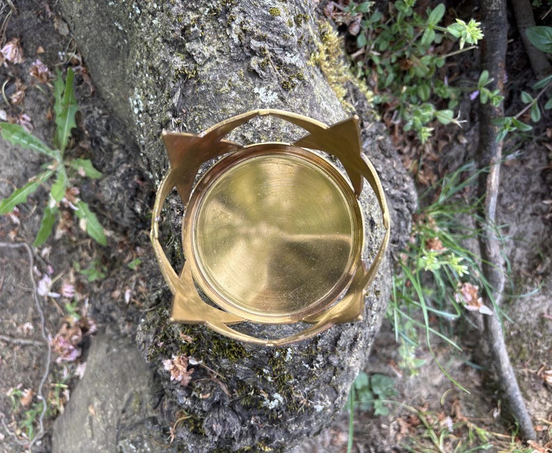 Puede incluir: Un portavelas de metal dorado con base circular y dise&ntilde;o calado. El portavelas tiene un patr&oacute;n de estrella en el borde superior. El portavelas est&aacute; sobre un tronco de &aacute;rbol.