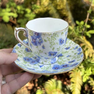 May include: A vintage teacup and saucer set. The white porcelain cup and saucer are decorated with blue floral patterns and gold trim. The cup has a curved handle. The saucer is round. The set is displayed outdoors.