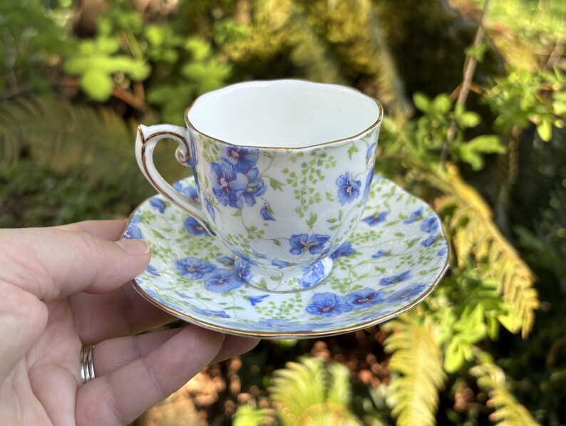 May include: A vintage teacup and saucer set. The white porcelain cup and saucer are decorated with blue floral patterns and gold trim. The cup has a curved handle. The saucer is round. The set is displayed outdoors.