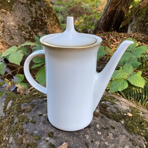 Może przedstawiać: Biały ceramiczny dzbanek do kawy z złotą obwódką wokół pokrywki i dzióbka. Dzbanek stoi na porośniętym mchem kamieniu.