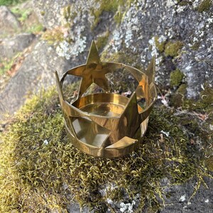 Puede incluir: Un portavelas de lat&oacute;n con dise&ntilde;o de estrella. El soporte tiene una base circular y est&aacute; rodeado de recortes en forma de estrella. El portavelas est&aacute; sobre una roca cubierta de musgo, con un fondo natural al aire libre.