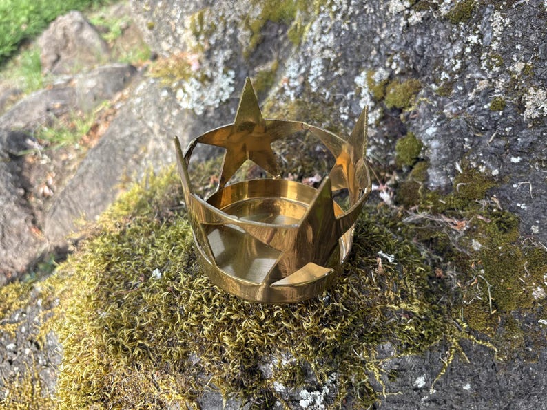 Puede incluir: Un portavelas de lat&oacute;n con dise&ntilde;o de estrella. El soporte tiene una base circular y est&aacute; rodeado de recortes en forma de estrella. El portavelas est&aacute; sobre una roca cubierta de musgo, con un fondo natural al aire libre.