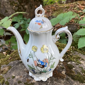 May include: White porcelain teapot with a gold trim and a floral design. The teapot features a small gnome-like figure swinging from a flower stem. The lid of the teapot has a blue bird design.