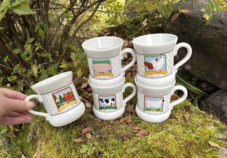May include: White ceramic mugs with farm animal illustrations. Each mug features a different animal: a pig, a cow, a sheep, and a chicken. The mugs have a patterned design and a handle. The mugs are stacked on top of each other.