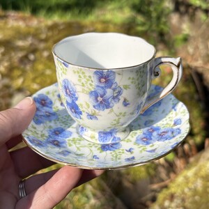 May include: A floral teacup and saucer set. The cup and saucer are white with a blue floral pattern and gold trim. The teacup has a curved handle. The saucer is round and has a matching floral design.