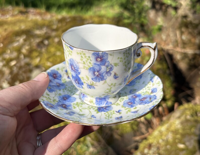 May include: A floral teacup and saucer set. The cup and saucer are white with a blue floral pattern and gold trim. The teacup has a curved handle. The saucer is round and has a matching floral design.