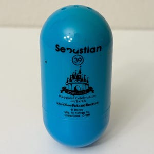 May include: A light blue, egg-shaped collectible featuring the name "Senustian" and the number "39". It has a castle graphic and text that reads "Happiest Celebration on Earth" and "2005-2006". The bottom of the collectible has the Disney Parks and Resorts logo.