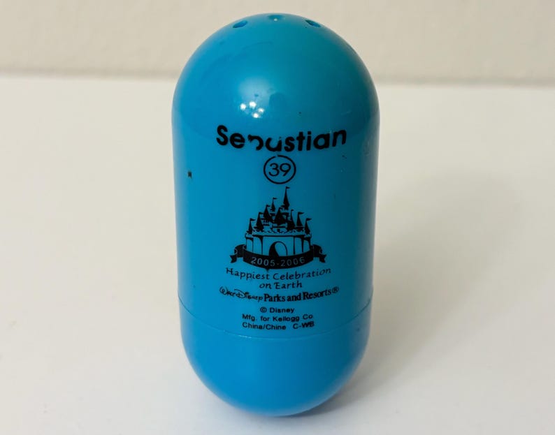 May include: A light blue, egg-shaped collectible featuring the name "Senustian" and the number "39". It has a castle graphic and text that reads "Happiest Celebration on Earth" and "2005-2006". The bottom of the collectible has the Disney Parks and Resorts logo.