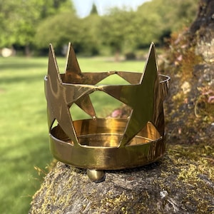 Puede incluir: Un portavelas de metal dorado con recortes de estrellas. El portavelas es circular con una base plana y un borde. Los recortes de estrellas est&aacute;n espaciados uniformemente. El portavelas est&aacute; sobre una superficie musgosa.