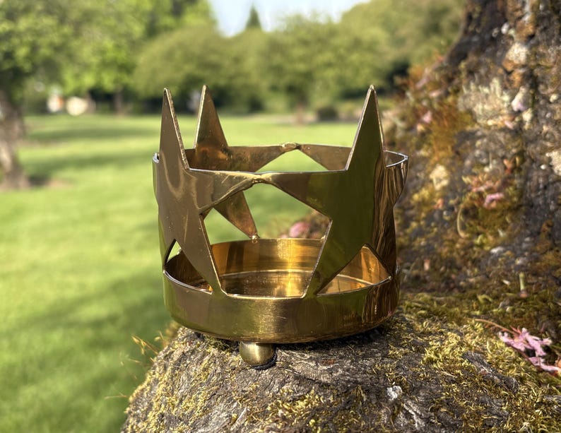 Puede incluir: Un portavelas de metal dorado con recortes de estrellas. El portavelas es circular con una base plana y un borde. Los recortes de estrellas est&aacute;n espaciados uniformemente. El portavelas est&aacute; sobre una superficie musgosa.
