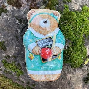 May include: A vintage bear-shaped tin with a teacher theme. The bear wears a teal dress with alphabet letters and holds a chalkboard that says "TO MY TEACHER" with a red apple. The tin is on a mossy rock.