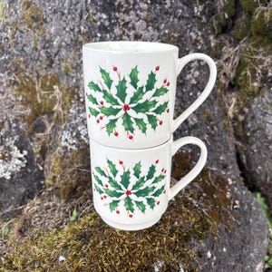 May include: Two stacked white ceramic mugs featuring a repeating green holly leaf and red berry pattern centered on the front. Each mug has a smooth cylindrical body, a curved handle on the right side, and a slightly tapered base for stacking.