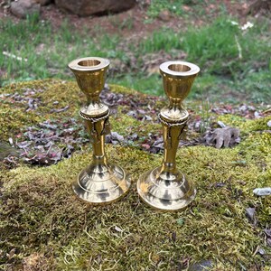 May include: Two brass candle holders with a detailed design. The candle holders have a round base, a decorative center, and a cup-shaped top. They are placed on a mossy rock with a blurred green background.