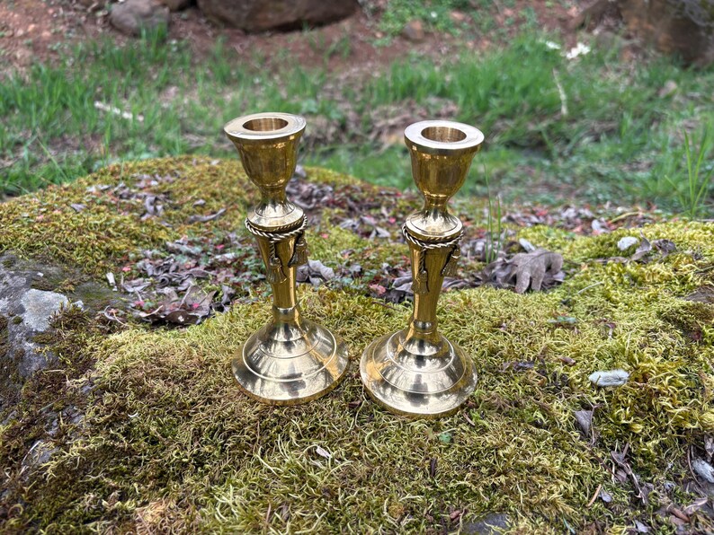 May include: Two brass candle holders with a detailed design. The candle holders have a round base, a decorative center, and a cup-shaped top. They are placed on a mossy rock with a blurred green background.
